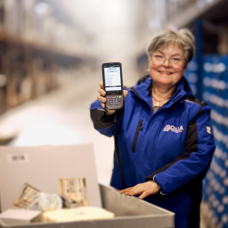 Mitarbeiterin von Cella im Lager zeigt Handy mit "E-Commerce in Emsbüren" und Optionen wie Kommissionierung, Wareneingang, Einlagerung und Inventur auf dem Bildschirm.